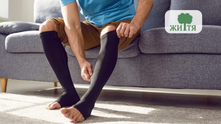 Person seated on a gray sofa, adjusts black knee-high compression socks on their legs. Brand logo appears in the corner.