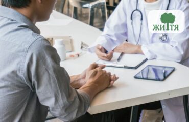 Patient sits across from a doctor at a clinic desk, hands folded, with a tablet and clipboard on the table; a tree logo appears in the top-right corner.