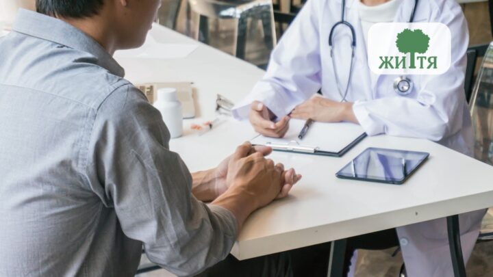 Patient sits across from a doctor at a clinic desk, hands folded, with a tablet and clipboard on the table; a tree logo appears in the top-right corner.